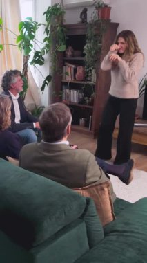 Genç bir kadın, sıcak bir oturma odasında arkadaşlarıyla tahmin oyunu oynarken rol yapıyor ve el kol hareketi yapıyor.