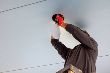 İşçi açık ve parlak alanda merdivenin üzerinde dururken tavan lambası armatürünü bağlar.