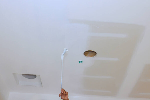 Painter is engaging in ceiling paints by reaching up with spray tool towards surface above.