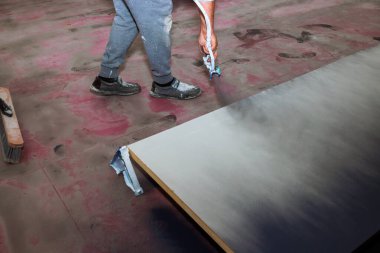 İşçi, tozlu zeminde dururken tahta tahtaya sprey tabanca sürüyor.