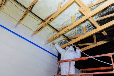 Koruyucu ekipmandaki ressam, inşaat alanının köpük tavan kirişlerine kırıcı boya yalıtımı uygulamak için sprey makinesi kullanıyor..