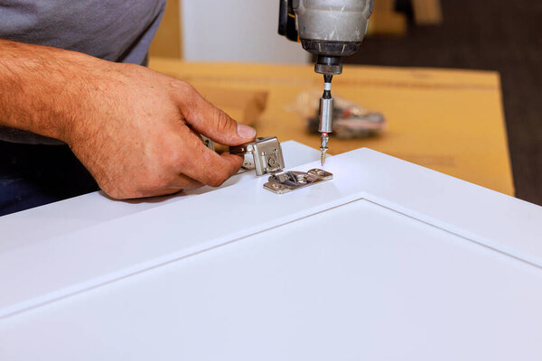 Работник прикрепляет петлю к белой двери шкафа, используя дрель, в то время как