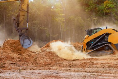 İki ağır makine, ağaçlarla çevrili inşaat alanındaki toprak kalıntılarını temizlemekle meşgul..