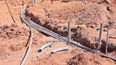 İşçiler inşaat sahasındaki kumla çevrili siperlere gri borular döşüyorlar.