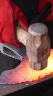 Bir zanaatkar, çekiç ve baltayla örs üzerinde kırmızı metali şekillendiriyor. Bu sırada usta demircilik yapıyor..