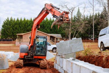 Bir kazıcı inşaat sahasında istinat duvarları inşa ederken yerleştirmek için beton blokları toplar