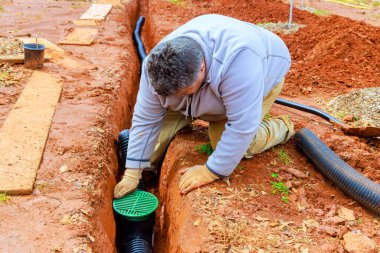 Adam kanalizasyon sistemi kurmak için bahçede çalışıyor. Siper kazıyor, toprak boruları dikiyor.