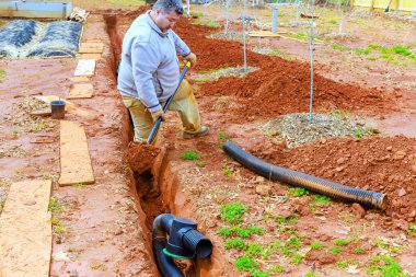 İşçi bahçe kazma siperlerinde çalışıyor, drenaj boruları kuruyor, toprak hazırlıyor.