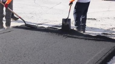İki işçi inşaat alanında yere taze asfalt serpmek için kürek ve tırmık kullanıyor.