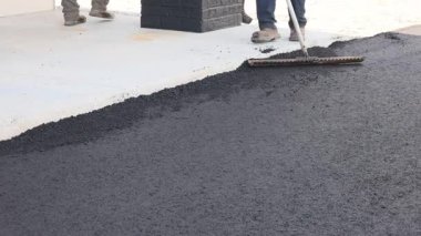 İşçiler evin yanındaki yol için pürüzsüz bir yüzey oluşturmak için tırmık kullanarak asfalt yayarlar..