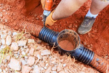 İşçiler kanalizasyon borularını tamir etmek için kazı yapıyorlar. Çamur, çakıl, emniyet eldiveni ve ayakkabı giyiyorlar..