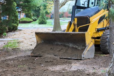 Sarı traktör, güneşli bir günde çimlerle çevrili bahçeyi kazıyor.