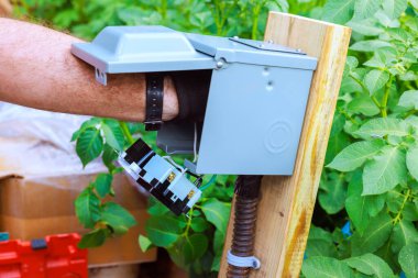 Yeşilliklerle çevrili gri kutuya elektrik parçaları yerleştirmek için ellerini kullanan kişi