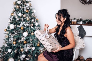 Esmer gülümseyen mutlu kadın hediye paketi açıyor, yeni yıl akşamı, Noel süslemeli ağaç. Yaşam tarzı ve alışveriş konsepti. Çift ırklı kız tebrikleri tatille karşılıyor.