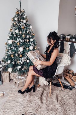Esmer gülümseyen mutlu kadın hediye paketi açıyor, yeni yıl akşamı, Noel süslemeli ağaç. Yaşam tarzı ve alışveriş konsepti. Çift ırklı kız tebrikleri tatille karşılıyor.