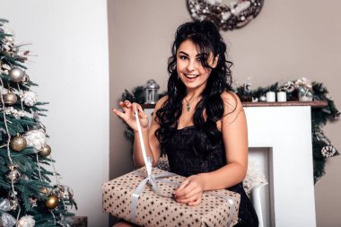 Esmer gülümseyen mutlu kadın hediye paketi açıyor, yeni yıl akşamı, Noel süslemeli ağaç. Yaşam tarzı ve alışveriş konsepti. Çift ırklı kız tebrikleri tatille karşılıyor.