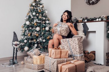 Esmer gülümseyen mutlu kadın hediye paketi açıyor, yeni yıl akşamı, Noel süslemeli ağaç. Yaşam tarzı ve alışveriş konsepti. Çift ırklı kız tebrikleri tatille karşılıyor.