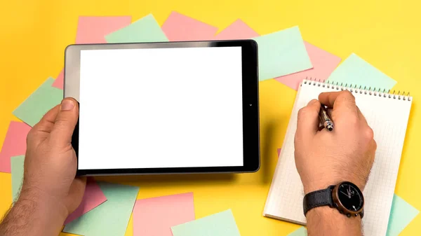 Cropped view mock up image black tablet pc white blank screen isolated yellow bright background. Colorful memo sticky pin clips empty notes scattered chaotic manner. Man writting in notepad with pen