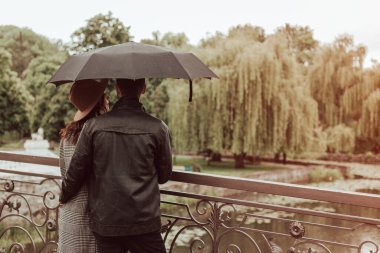 Heteroseksüel çiftin, kara şemsiyeli, yağmurlu ve kasvetli bir havada nehir kenarındaki köprüde duruş görüntüsü. Kopyalama alanı ve bokeh arkaplanı olan resim.