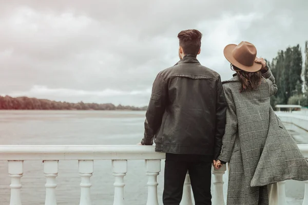 Back view of loving couple standing on the pier on lake or river in cloudy windy weather. Brunette lover and lady in hat spending time together. Image with copy space and blur background.