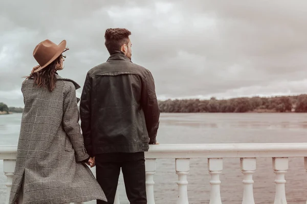 Back view of loving couple standing on the pier on lake or river in cloudy windy weather. Brunette lover and lady in hat spending time together. Image with copy space and blur background.