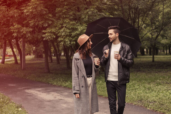 Young couple walking in the spring green city park, drinking hot coffee paper eco recycle cup. Two lovers husband wife spending time together, discuss news with black umbrella to shelter the rain.