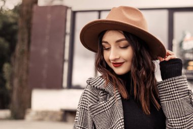 Caddedeki kafe restoranının önünde dikilen çekici kadını yakın plan çek. Kahverengi şapkalı, gri ceketli, polonyalı siyah kazaklı, başka tarafa bakan bir kadın. Kız vaktini sonbaharda ya da ilkbaharda, soğuk havada temiz havada geçirir..