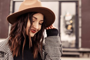 Caddedeki kafe restoranının önünde dikilen çekici kadını yakın plan çek. Kahverengi şapkalı, gri ceketli, polonyalı siyah kazaklı, başka tarafa bakan bir kadın. Kız vaktini sonbaharda ya da ilkbaharda, soğuk havada temiz havada geçirir..