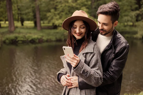 Young couple spring city park, using holding looking at cell mobile phone modern gadgets. Two lovers newlyweds husband wife spending time together, hugging and kissing. People lifestyle concept.