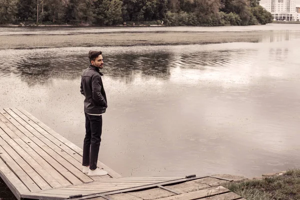 全长的肖像画 严肃体贴的男人站在河边的木制码头上 闷闷不乐的下雨天 望着远方 穿着黑色皮革保暖夹克 浑浊的水流自然 — 图库照片