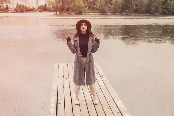 Full length portrait of woman standing on the wooden pier near river lake moody rainy weather, looking at the camera. Dressed warm gray coat poloneck black sweater. Turbid water flows nature