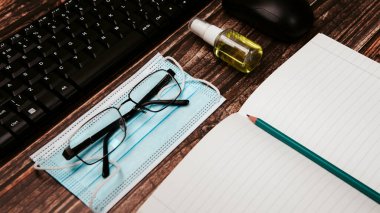 Masasında mavi yüz maskeleri olan küçük bir şişe antiseptik el dezenfektanı, koronavirüs önleme. Klavye ve fare gözlükleri. Ev konseptinde çalışma, kalem defteri.