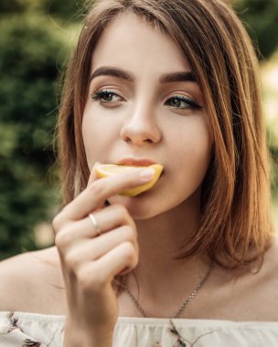 Beyaz bahar elbisesi giymiş uzun saçlı bir kadının portresi. Yaz parkında duruyor ve bir dilim sulu limon yiyor. Fotokopi alanı ve doğa arka planı olan bir resim..