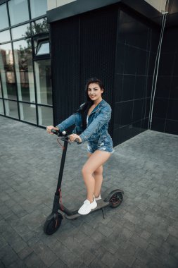 Şehrin caddesinde duran elektrikli scooterlı kadının dikey portresi. self-servis sokak taşımacılık kiralama servisi. Akıllı telefonlu kentsel çevre dostu enerji aracı kiralayın. 