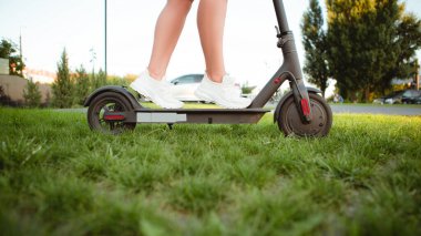 Elektrikli scooter 'ı olan bir kadının şehir parkında yeşil çimen ya da çimenlikte duruşu. self-servis sokak taşımacılık kiralama servisi. Kentsel çevre dostu enerji aracı kirala.