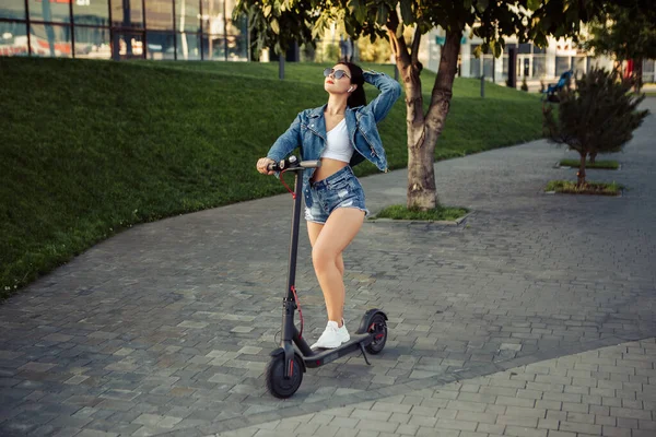 Şehrin caddesinde duran elektrikli scooterlı kadın portresi. self-servis sokak taşımacılık kiralama servisi. Akıllı telefon uygulaması olan kentsel çevre dostu enerji aracı kiralayın. 