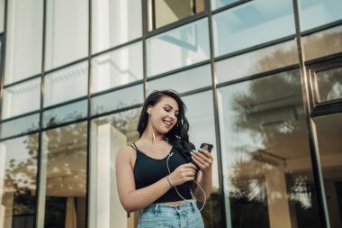 Mobil cihazlı, beyaz kulaklıklı, dışarıda müzik dinleyen gülümseyen kadın portresi. Kopyalama alanı olan resim.