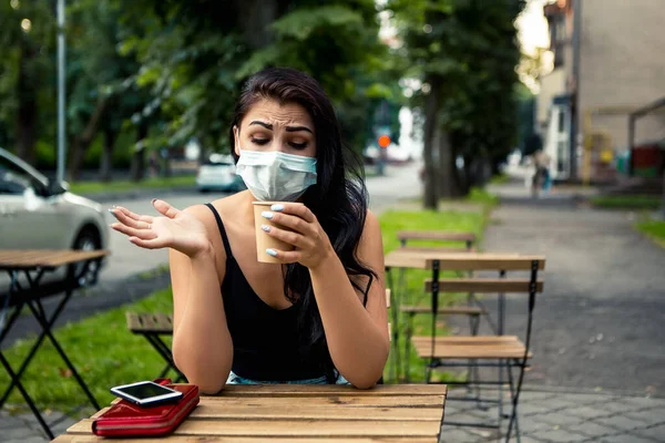 Young confused woman sitting outside near street cafe or restaurant small business with hot coffee beverage, coronavirus pandemic. Lady wear blue face disposable mask, protect from disease pneumonia.