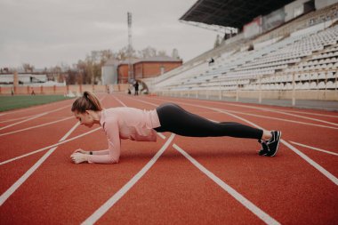 Kız, stadyumun koşu bandında kalas yapıyor. Bulanık arka planda eğitim gören insanlar. Sportif kadın dışarıda spor yapıyor. Boş alanı kopyala. Güçlü kadın pozisyon aldı..
