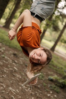 Şehir parkında baş aşağı asılı duran ve elinde beyaz tabletle gezen çekici bir kızın dikey portresi. Gülümseyen kıvırcık kadın dışarıda eğleniyor ve 5G ağıyla cihaz kullanıyor..