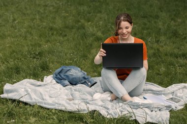 Ekoseli dizüstü bilgisayarı olan mutlu genç bayan dizüstü bilgisayarla çalışıyor. Turuncu tişörtlü kızın neşeli bir gülümsemesi var. Dışarıdaki dişlek öğrenci. Boşluğu kopyala.