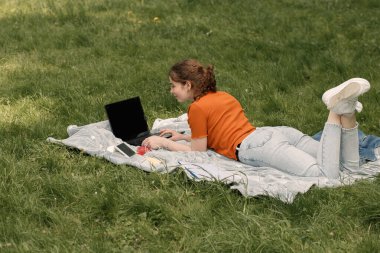 Şehir parkında ya da avluda yeşil çimlerde yatan ve dizüstü bilgisayar kullanarak yazı yazan çekici kız. Dışarıda serbest çalışan bir kadın. Teknolijiyi kullanan kadın.