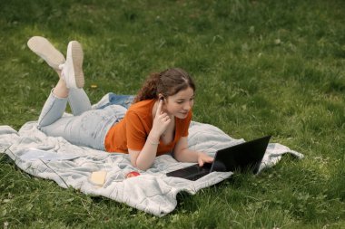 Şehir parkında yeşil çimlerin üzerinde yatan çekici genç bir kızın yatay portresi. Bahçede kablosuz kulaklıkları ve dizüstü bilgisayarı olan neşeli bir kadın..