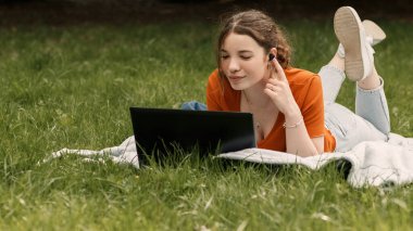 Yeşil çimenlerin üzerinde dizüstü bilgisayarı ve kablosuz kulaklığı olan güzel, kıvırcık bir kızın portresi. Çağdaş teknolojileri kullanan bir kadın. Kopyalama alanı ile uzaklık çalışması.