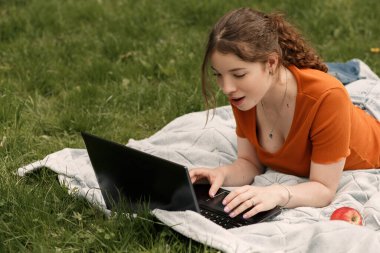 Vay canına! İnternette sosyal ağlarda sohbet eden şaşırmış genç kız öğrencinin portresi. Yeşil çimlerde dizüstü bilgisayarı ve kırmızı elması olan bir kadın. Boşluğu kopyala.