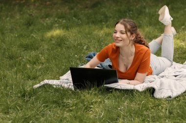Genç gülümseyen çekici bir üniversite öğrencisinin portresi şehir parkındaki çimlerde ya da çimlerde yatar ve bir şeye bakar. Dizüstü bilgisayarı olan güzel kız. Kopyalama alanı olan resim