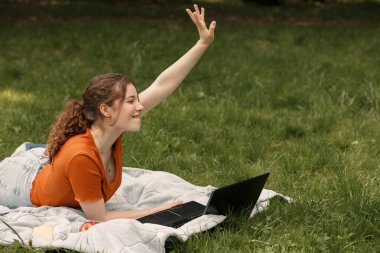 Genç gülümseyen mutlu üniversite öğrencisinin hayat tarzı portresi çimlerde yatıp dizüstü bilgisayarla çalışırken arkadaşlarına el sallıyor. Şehir parkında fotokopi alanı olan bir kadın.