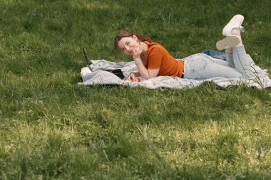 Kameraya bakan genç ve güzel bir kızın yatay hayat portresi. Dizüstü bilgisayarı olan güzel bir kadın, şehir parkında ya da bahçesinde, metin boşluğu olan çimlerde yatıyor..