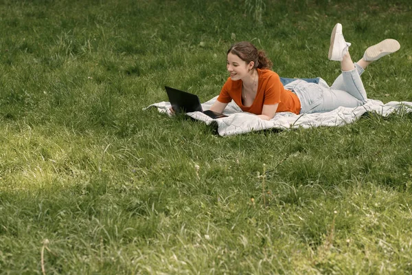 Genç tatlı kıvırcık kız kolej öğrencisi dizüstü bilgisayarla çimlerin üzerinde yatıyor ve bulutlu bir havada web sitesinde bilgi arıyor. Belgelere metin yazan kadın.