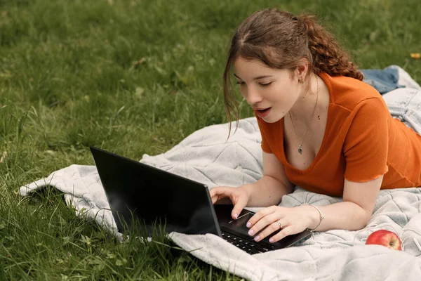 Vay canına! İnternette sosyal ağlarda sohbet eden şaşırmış genç kız öğrencinin portresi. Yeşil çimlerde dizüstü bilgisayarı ve kırmızı elması olan bir kadın. Boşluğu kopyala.